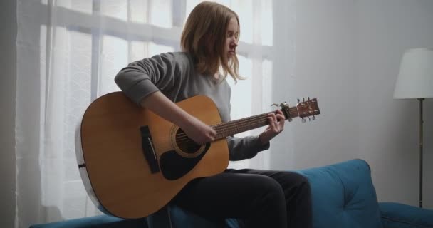 A lány gitározik a kanapén. Egy nő egyedül szomorú zenét kreál egy fehér nappaliban. A művész akusztikus gitáron játszik. A zenész komponál egy dallamot. - Felvétel, videó