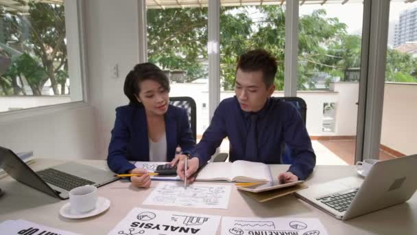 Medium shot of young Asian businessman and businesswoman sitting together at working table, looking at papers with schemes and chats, talking and making some calculations - Footage, Video