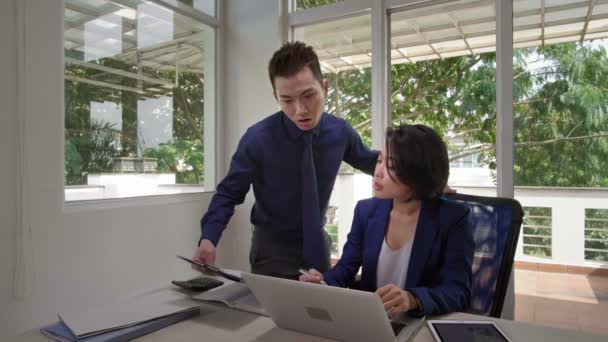 Medium shot van Aziatische zakenvrouw zitten voor laptop en Aziatische zakenman staan achter haar, met documenten in zijn handen, wijzen met zijn vinger op een aantal regelingen en praten met haar - Video