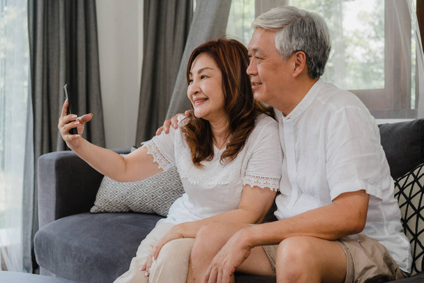 アジア系のシニアカップルのビデオ通話。中国人のアジア系高齢者の祖父母は、自宅のコンセプトでソファの上に横たわっている間、家族の孫と話す携帯電話のビデオ通話を使用して. - 写真・画像