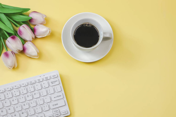Horní pohled nebo ploché položení počítačové klávesnice, fialová bílá tulipánová kytice a šálek kávy na žlutém pozadí s kopírovacím prostorem. - Fotografie, Obrázek