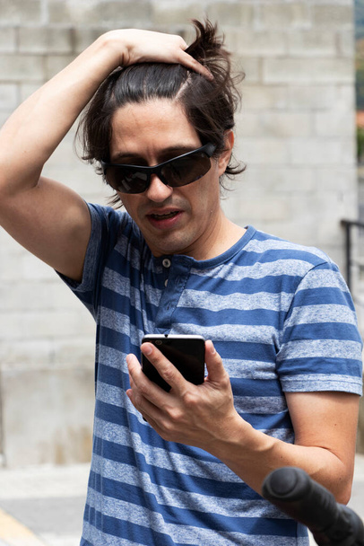 Black haired man in blue striped shirt holds cell phone removing hair from face with worried expression - Φωτογραφία, εικόνα
