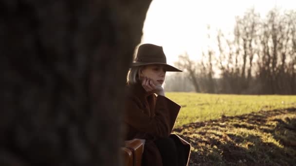 fille élégante dans un chapeau avec une valise assise sur l'herbe près du champ au coucher du soleil - Séquence, vidéo