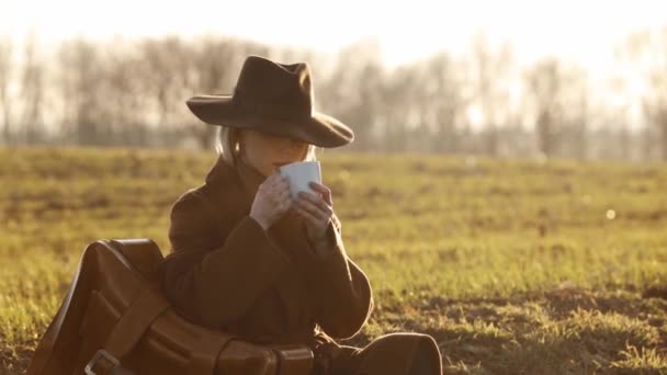 stijlvol meisje in een hoed met een koffer en een kopje koffie zittend op het gras bij het veld bij zonsondergang - Video