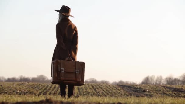 стильна дівчина в капелюсі з валізою, що йде в полі на заході сонця
 - Кадри, відео