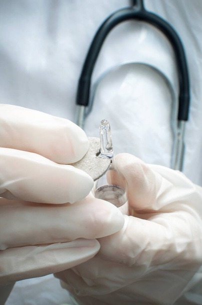 Une femme médecin en gants médicaux ouvre une ampoule en verre avec le médicament. Mains en gros plan, verticales. Concentration sélective. - Photo, image