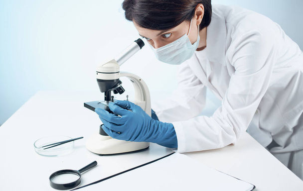 medico donna in abito medico e guanti guardando attraverso un microscopio su un tavolo all'interno - Foto, immagini