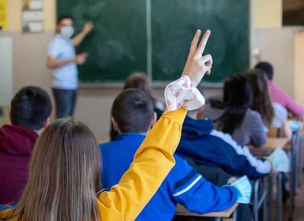 Вид спереду шкільної дівчини з піднятою рукою з маскою безпеки в класі, вчитель на задньому плані. Концепція захисту пандемії в освіті
 - Фото, зображення