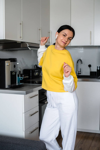 Brunette girl with bluetooth headphones dances in the kitchen in the morning and makes coffee in a coffee machine - Foto, afbeelding