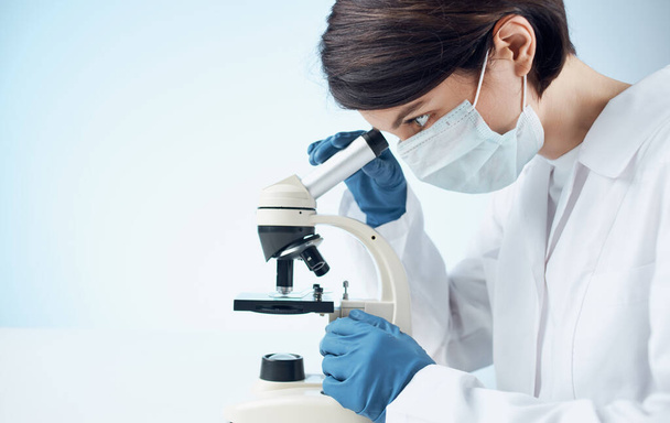 vrouw arts in een medische jurk en handschoenen kijken door een microscoop op een tafel binnen - Foto, afbeelding