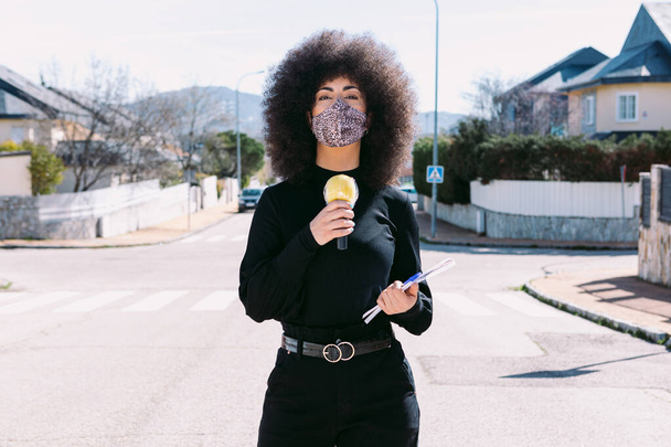 Afro saçlı, kendini virüsten korumak için leopar derisi maske takan bir kadın televizyon muhabiri sokakta bir haber yapıyor. - Fotoğraf, Görsel