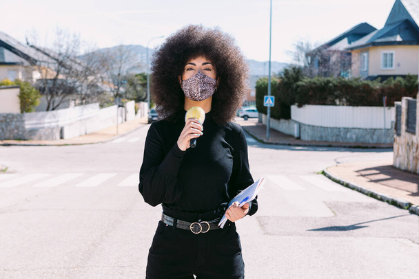 Afro saçlı, kendini virüsten korumak için leopar derisi maske takan bir kadın televizyon muhabiri sokakta bir haber yapıyor. - Fotoğraf, Görsel
