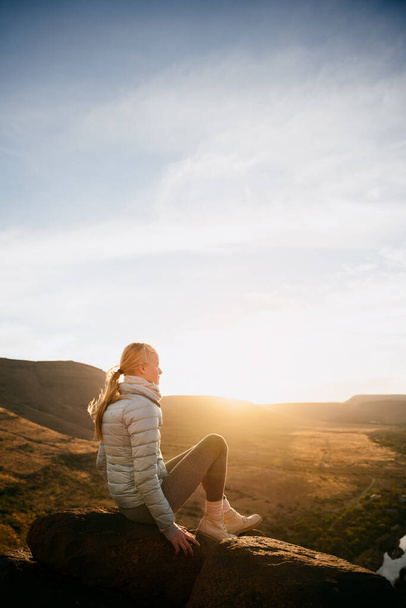 Joven caucásico adolescente sentado en la parte superior de la roca viendo puesta de sol senderismo exuberante montaña  - Foto, imagen