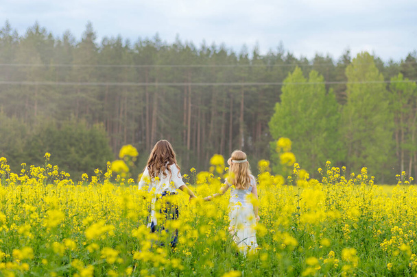 Dos chicas están caminando a lo largo de un campo de colza en flor. - Foto, imagen