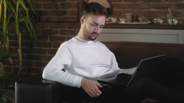 Fiatal vonzó férfi ül egy kanapén egy hangulatos otthoni környezetben egy fekete macska és gépelés egy laptop. Távoli munka háziállatokkal. Egy karantén alá helyezett szabadúszó dolgozik otthon.. - Felvétel, videó