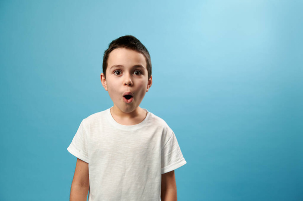 Geschokt jongen poseren met geopende mond uiten verrassing op blauwe achtergrond met kopieerruimte - Foto, afbeelding