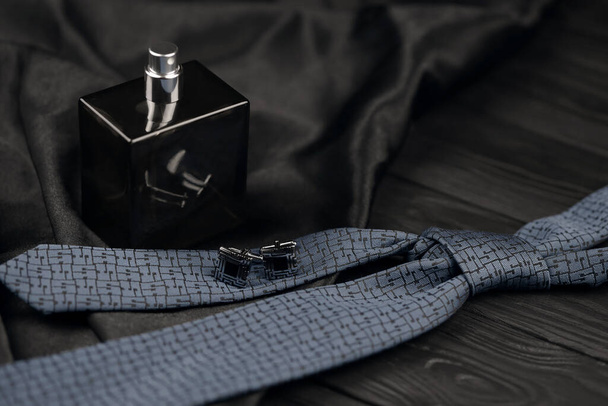 A bottle of mens cologne and cufflinks with blue tie lie on a black luxury fabric background on a wooden table. Mens classic accessories. Shallow DOF - Photo, Image