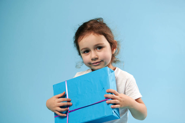 Αξιολάτρευτο κορίτσι με μπλε δώρο σε μπλε φόντο. Αντιγραφή χώρου. Ημέρα των παιδιών. Έννοια γενεθλίων. - Φωτογραφία, εικόνα