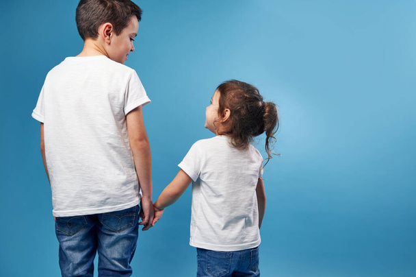 A boy and a girl stand with their backs to the camera on a blue background and look at each other holding hands tight. Copy space. - Photo, Image
