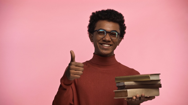 cheerful african american teenage boy in glasses holding books while showing thumb up isolated on pink - 写真・画像