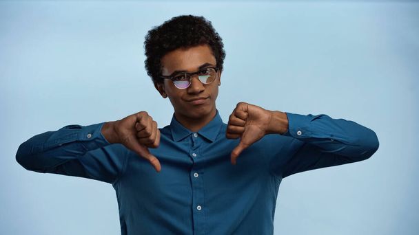 afroamericano adolescente en gafas mostrando pulgares abajo aislado en azul - Foto, Imagen