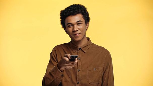 χαρούμενο Αφροαμερικανό έφηβο αγόρι κρατώντας τηλεχειριστήριο απομονωμένο στο κίτρινο - Φωτογραφία, εικόνα