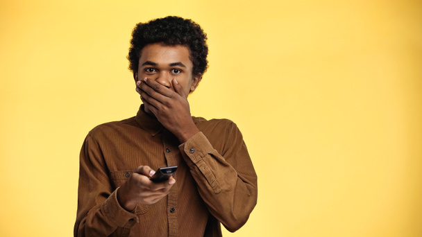 rizado africano americano adolescente cubierta boca mientras sosteniendo mando a distancia aislado en amarillo - Foto, Imagen