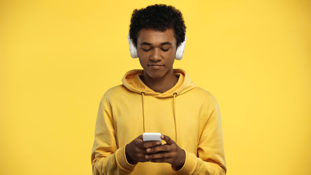 Afrykański amerykański nastolatek w słuchawkach bezprzewodowych przy użyciu smartfona izolowanego na żółto - Zdjęcie, obraz