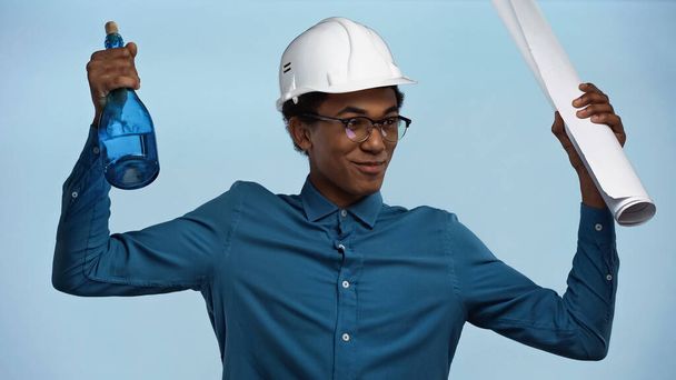 african american teenage boy in hard hat holding paper roll and bottle with champagne isolated on blue - Photo, Image