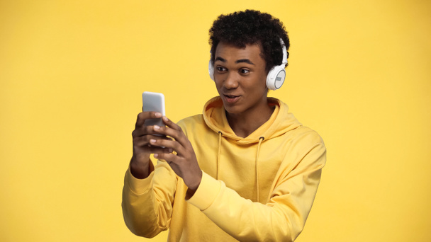 vrolijk afrikaanse tiener jongen in draadloze hoofdtelefoon met behulp van smartphone geïsoleerd op geel - Foto, afbeelding