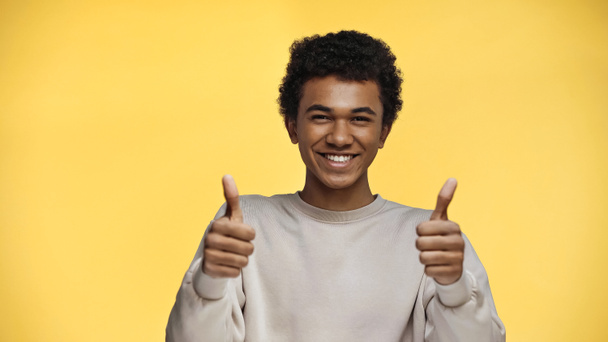 feliz afroamericano adolescente en sudadera mostrando pulgares hacia arriba aislado en amarillo - Foto, imagen