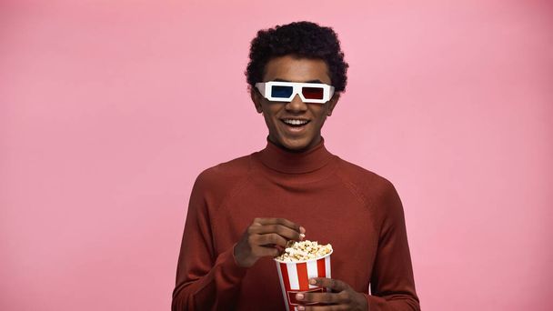happy african american teenager in 3d glasses holding popcorn bucket isolated on pink - Photo, Image