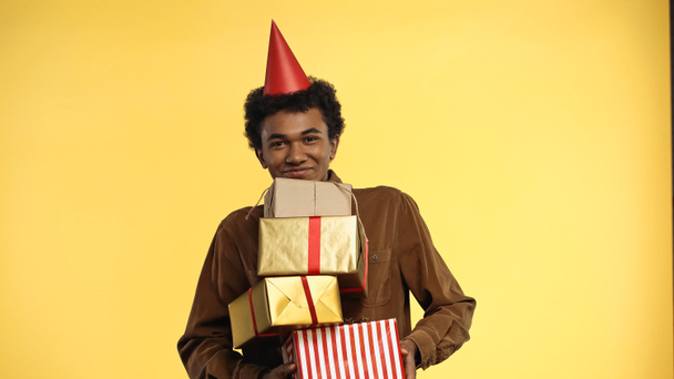 sonriente afroamericano adolescente en partido tapa celebración regalo cajas aislado en amarillo - Foto, imagen