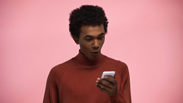 africký americký teenager v roláku svetr při pohledu na smartphone izolované na růžové - Fotografie, Obrázek