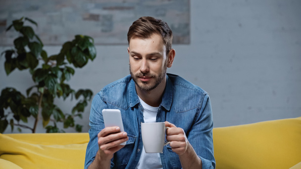 homem barbudo usando smartphone e segurando xícara de café na sala de estar  - Foto, Imagem