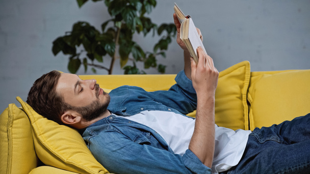 bearded man lying on sofa and reading book in living room - Photo, Image