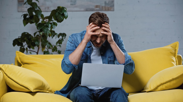 freelancer molesto sentado en el sofá y tocando la cabeza mientras usa el ordenador portátil - Foto, imagen