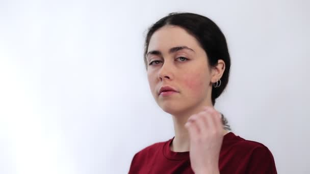 Portret van een jonge droevige vrouw die roodheid op haar wangen toont met haar hand. Witte achtergrond. Het concept van rosacea en couperose. - Video