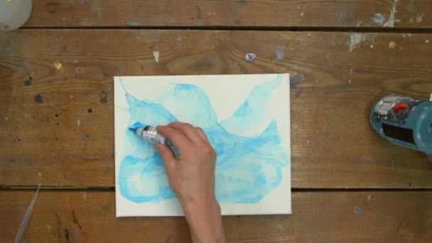 Vista superior de la artista pinta un cuadro abstracto, pinta formas abstractas con pintura azul sobre lienzo húmedo y utiliza pincel para distribuirlo - Metraje, vídeo