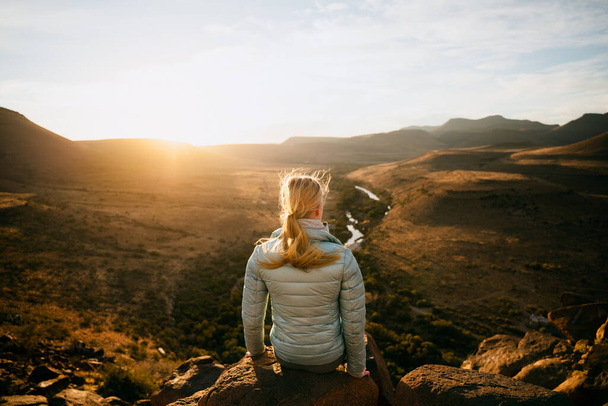 Νεαρή έφηβη κοπέλα κάθεται σε βράχο με θέα την πράσινη γη στο ηλιοβασίλεμα μετά από επιτυχημένη πεζοπορία. - Φωτογραφία, εικόνα