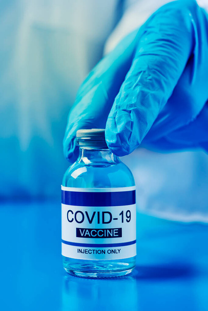 Mavi ameliyat eldiveni giymiş, mavi bir yüzey üzerinde simüle edilmiş kovid 19 aşı şişesini gösteren bir sağlık görevlisinin yakın çekimi. - Fotoğraf, Görsel