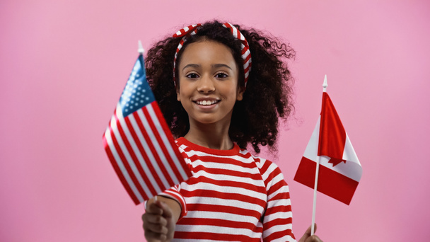 heureuse fille afro-américaine tenant des drapeaux d'Amérique et du canada isolés en rose  - Photo, image