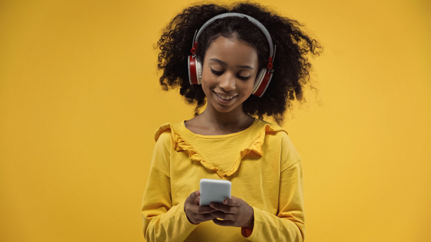 chica americana africana rizada en auriculares usando teléfono inteligente aislado en amarillo - Foto, Imagen