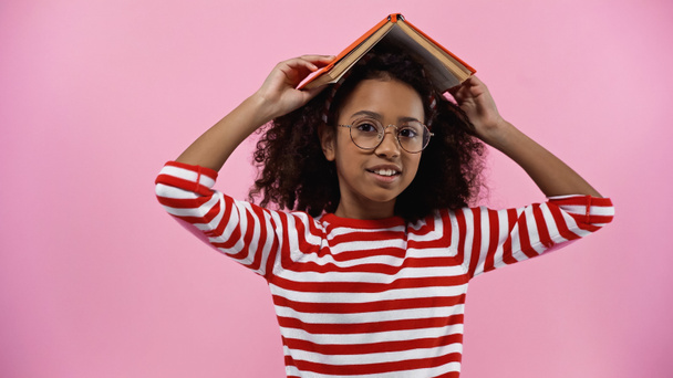 frisée afro-américaine fille dans des lunettes tenant livre au-dessus de la tête isolé sur rose  - Photo, image
