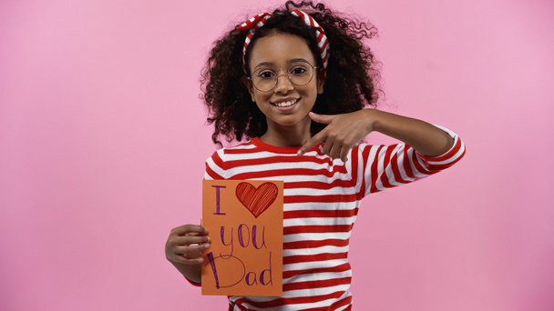 heureux afro-américaine fille dans des lunettes pointant du doigt à la carte de vœux avec je t'aime papa lettrage isolé sur rose - Photo, image