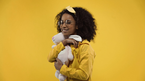 glücklich afrikanisch-amerikanische Mädchen in Brille hält weiche Häschen isoliert auf gelb - Foto, Bild