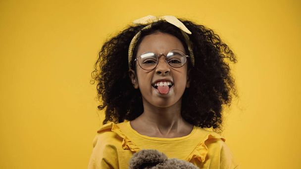 кудрявая африканская американка в очках, торчащая языком, изолированным на желтом - Фото, изображение