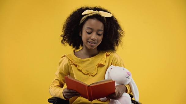 feliz africano americano chica con suave juguete lectura libro aislado en amarillo - Foto, Imagen
