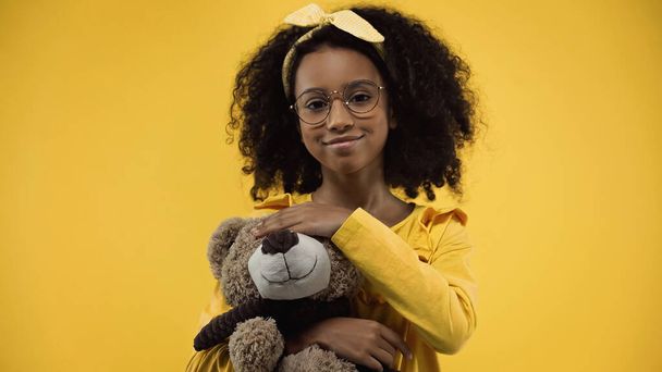 positive african american girl in glasses holding teddy bear isolated on yellow - Valokuva, kuva