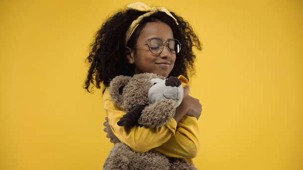 frisée afro-américaine fille dans des lunettes étreignant ours en peluche isolé sur jaune - Photo, image